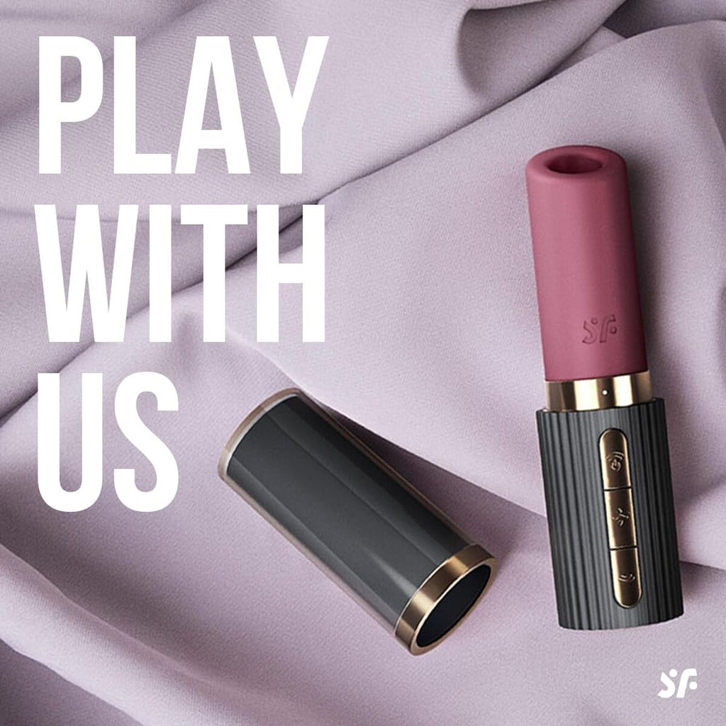 Pink lipstick vibrator with gold accents on a light purple fabric background, featuring the text 'Play With Us' and a logo.