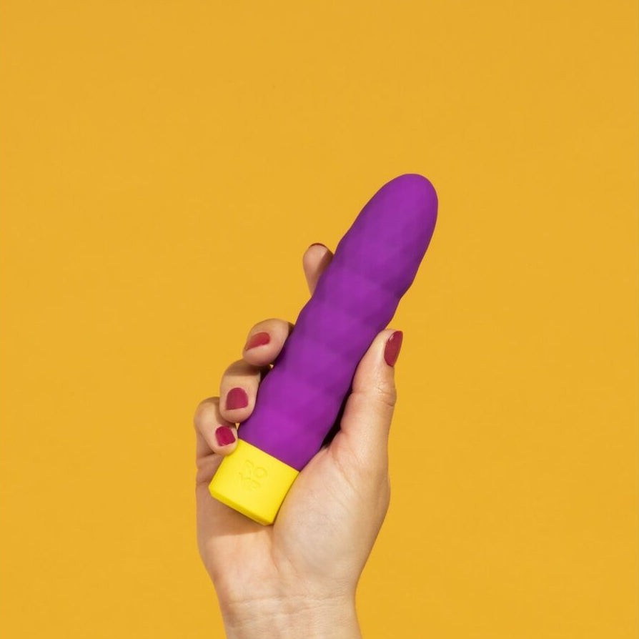 Purple and yellow vibrator adult toy held in a hand against a mustard yellow background
