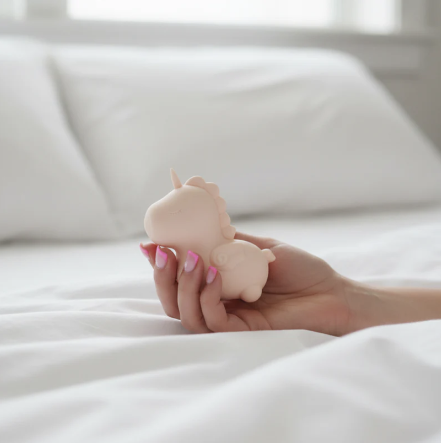 Woman hand holding a Pink unicorn-shaped sexual wellness device intended for adults only on white bedsheets